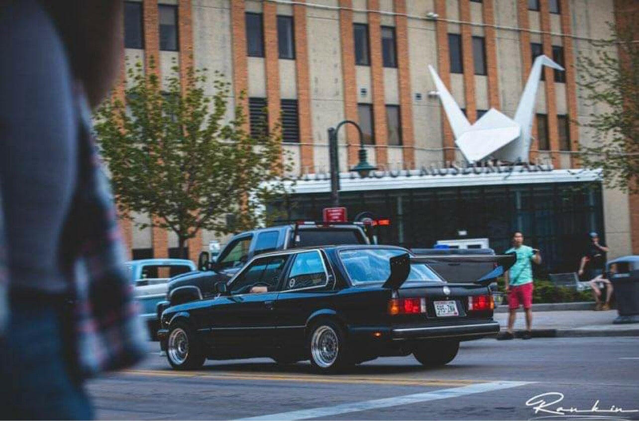 A classic black sedan, specifically the Street Wang 82-92 BMW 318 325 E30 by 9livesracing, drives in front of a building adorned with a large white origami crane art installation on its facade. Trees line both sides of the street, and a few pedestrians can be seen in the background, evoking the sleek blend often achieved by performance automotive parts.