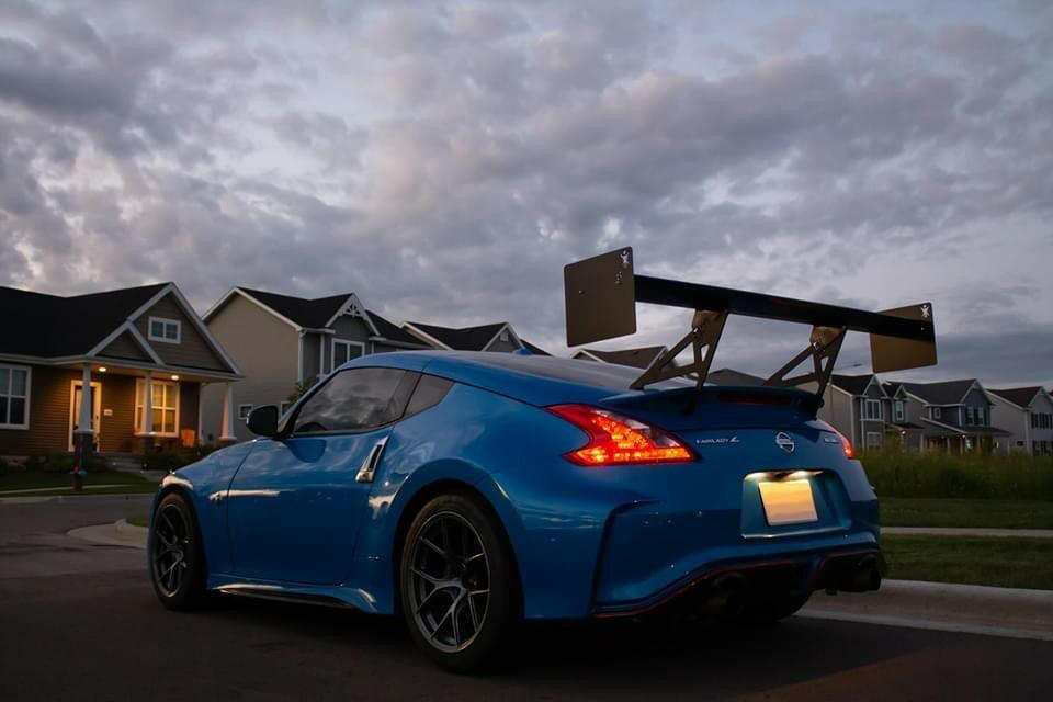 A sleek blue sports car, featuring the 370z Big Wang Kit '08-20 Z34 by 9livesracing with extruded aluminum wings for optimal down-force adjustments, is parked on a suburban street in front of houses. The sky shows a mix of clouds with a hint of sunset, creating a dramatic backdrop. The car's license plate is illuminated in the evening light.