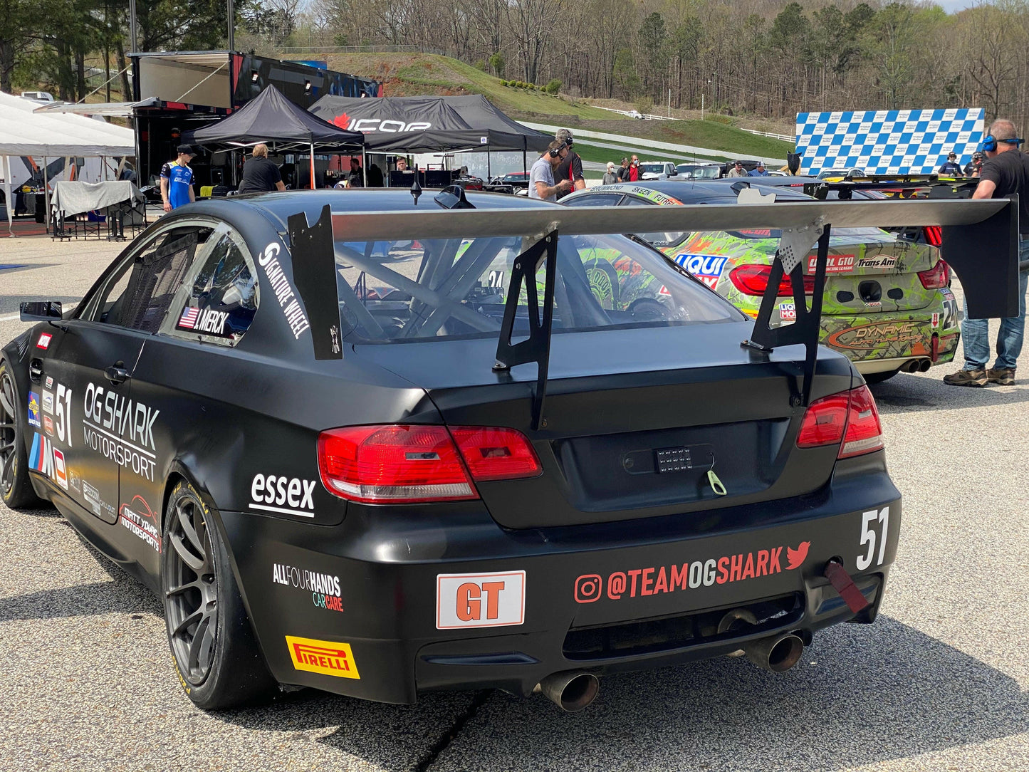 A sleek black race car numbered 51, adorned with various sponsor logos on the back, is parked in a racing area. Its rear wing features extruded aluminum wings from 9livesracing for increased downforce, and the exhaust system is visible. In the background, people stand near another race car equipped with a 3-Series Big Wang kit ’04-13 E90/E92/E93 by 9livesracing and some race equipment.