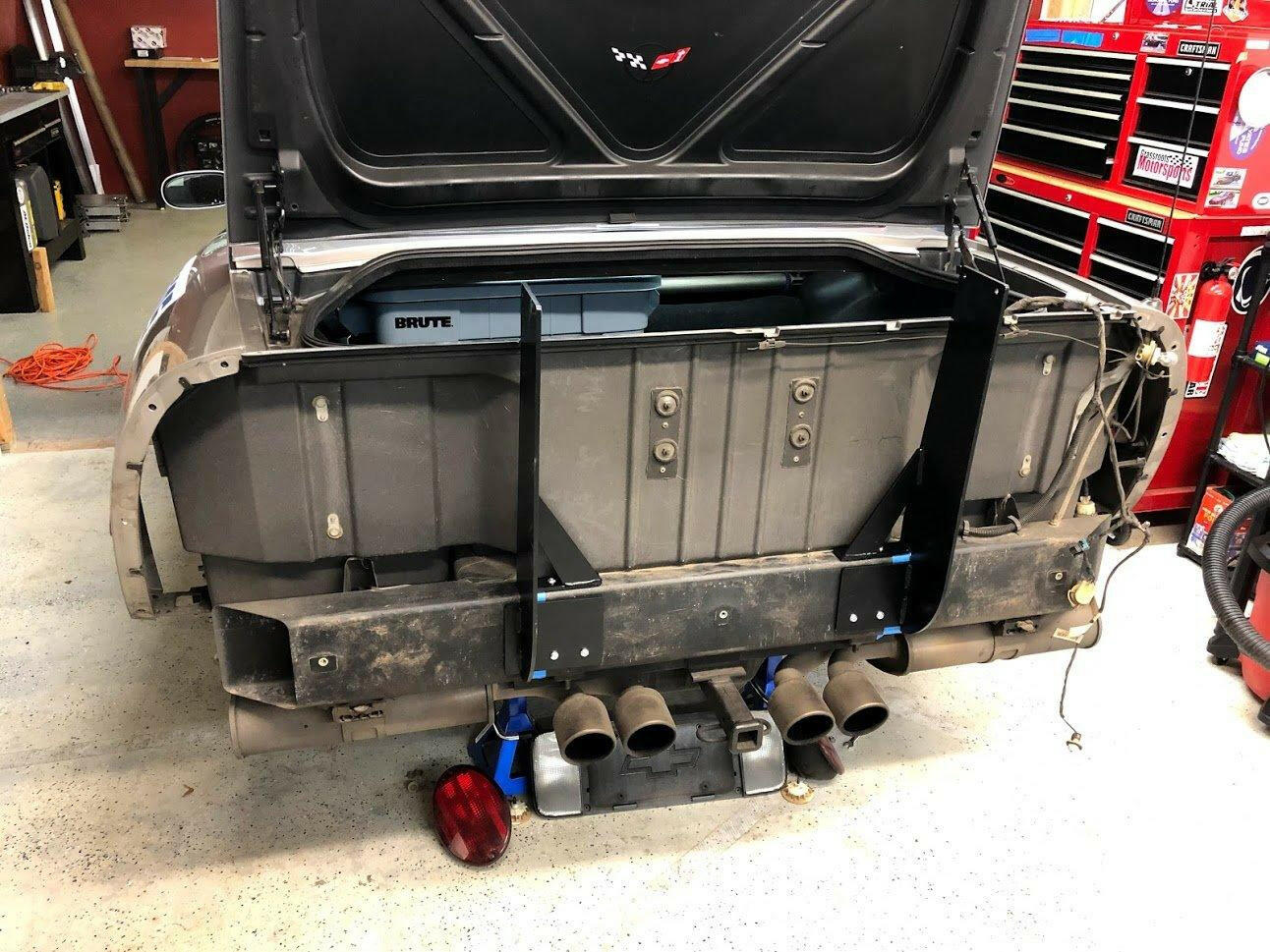 A Corvette with its rear bumper removed is shown in a garage, revealing a Big Wang GT3 dual exhaust system equipped with a tow hook. Various tools and equipment are visible in the background, while the open trunk displays a storage container labeled "BRUTE." This setup prominently features the Corvette Big Wang Chassis Mount ’97-13 C5/6 by 9livesracing.