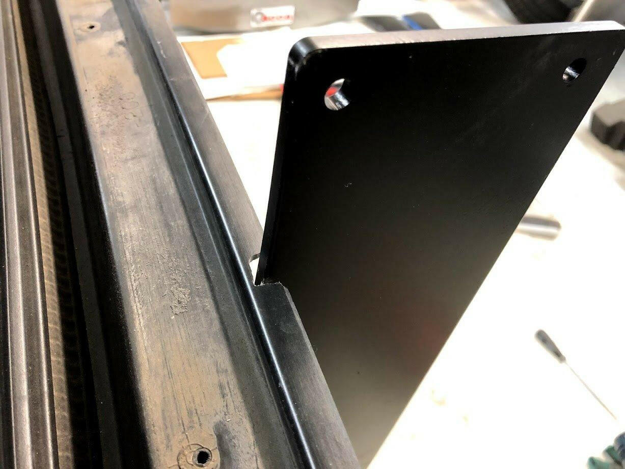 Close-up of a black metal bracket attached to a somewhat worn black metal frame. The bracket, which has two holes near the top edge and is positioned vertically, is part of the 9livesracing Corvette Medium Downforce Kit '97-04 C5. Some tools and other parts of the Aerodynamic kit are visible in the blurred background.
