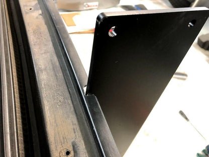 Close-up of a black metal bracket attached to a somewhat worn black metal frame. The bracket, which has two holes near the top edge and is positioned vertically, is part of the 9livesracing Corvette Medium Downforce Kit '97-04 C5. Some tools and other parts of the Aerodynamic kit are visible in the blurred background.