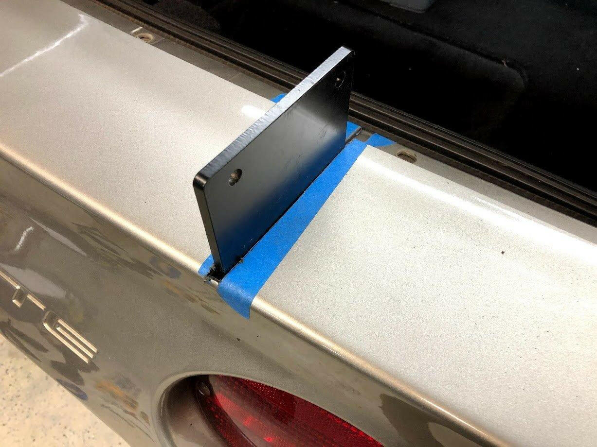 A black metal bracket from the "Corvette Medium Downforce Kit ‘97-04 C5" by 9livesracing is being trial-fitted onto the trunk lid of a silver C5 Corvette, secured temporarily in place with blue painter's tape. The bracket's positioning suggests it is part of an aerodynamic kit aimed at increasing downforce. The car's taillight is partially visible.