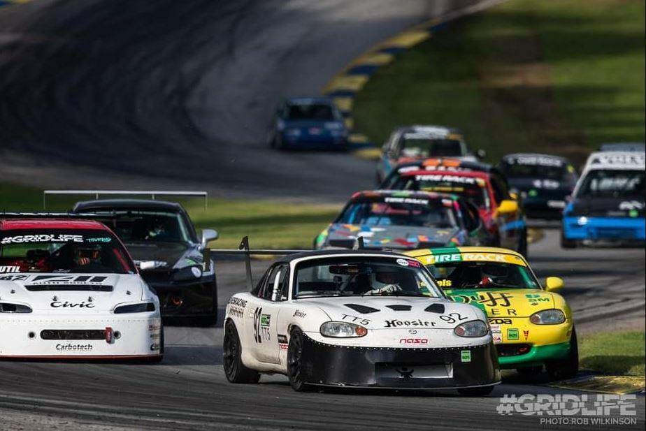 Several race cars rounding a corner on a racetrack, featuring an NB Miata fitted with the 9livesracing Miata Medium Downforce Kit '90-05 NA/NB in the forefront, closely followed by other brightly colored cars. Trees and a curving track are visible in the background. The image has the hashtag #GRIDLIFE and includes the photographer's name.