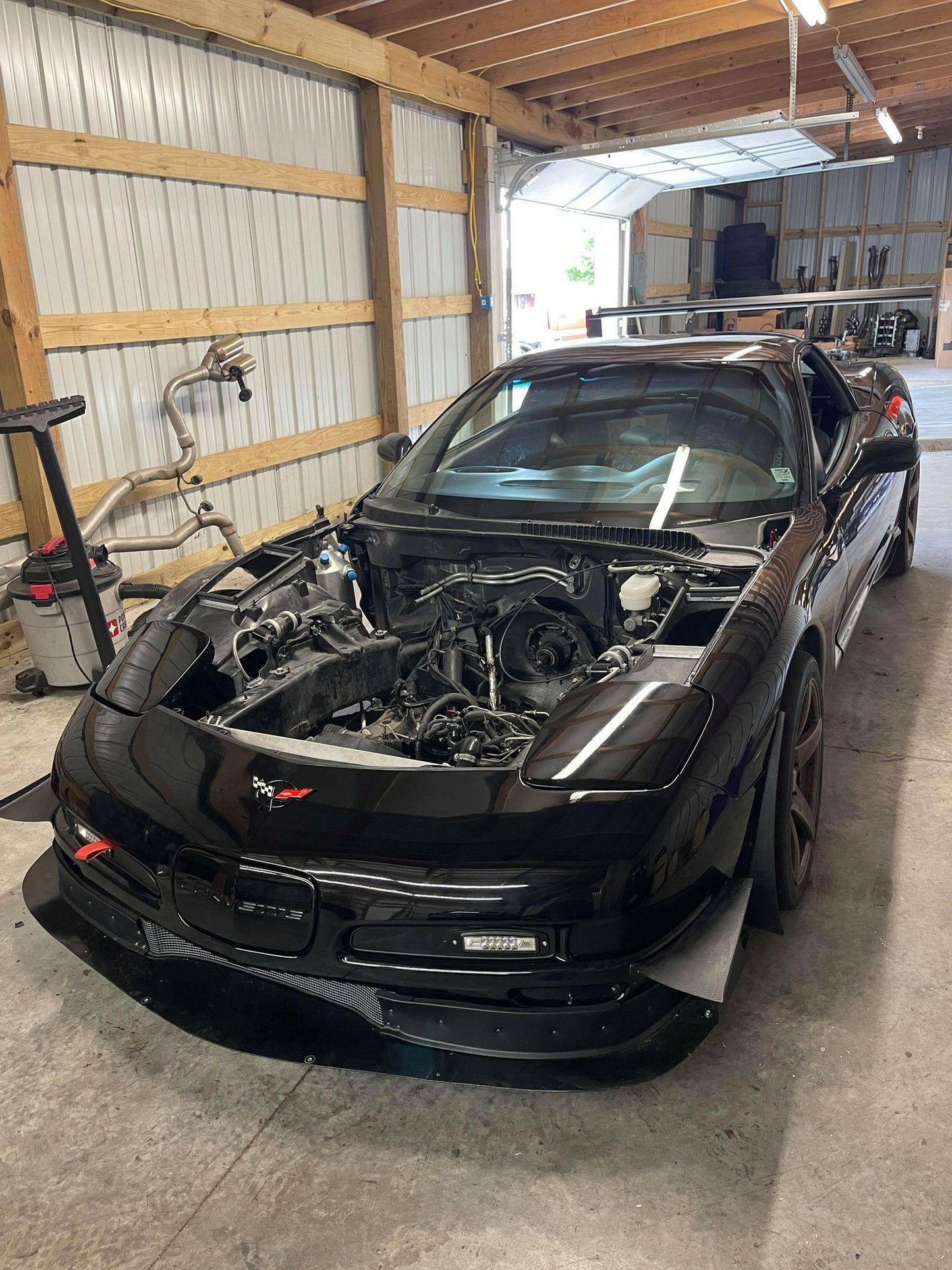 A black sports car, partially disassembled, is parked in a spacious garage with a wooden ceiling and metal walls. The hood is open and the engine appears to be removed. Various automotive tools and Nine Lives Racing C5 Corvette Car-Nards are scattered around the workspace.