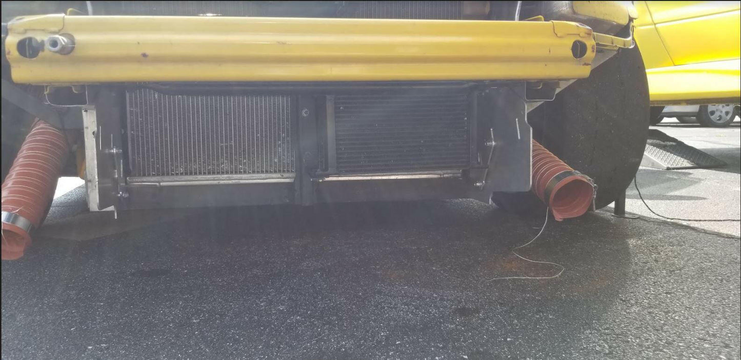 A close-up view of the underside of a yellow vehicle, fitted with the S2000 Sturdy Boii Splitter Mounts from 9livesracing, showcasing the radiator and various mechanical components, including meticulous radiator ducting. Two orange air ducts lead from the sides towards the radiator. Another yellow vehicle is visible in the background, resting on asphalt.