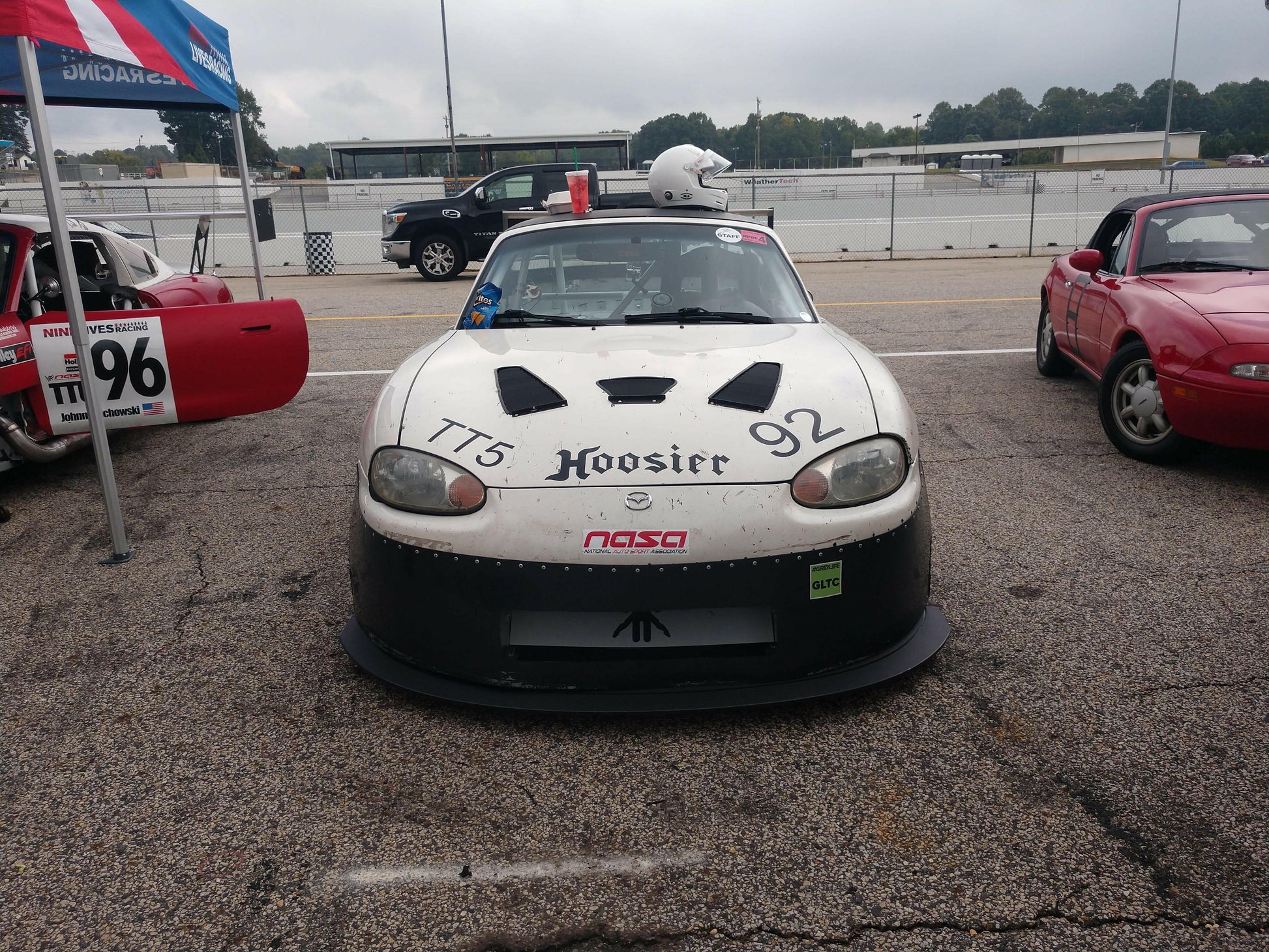 A racing car featuring a sleek white and black design and the number 92 alongside "Hoosier" on the hood is parked on a race track, fitted with the Miata Medium Downforce Kit '90-05 NA/NB from 9livesracing. The driver’s side door displays the number 96, and there is a helmet resting atop the car while other race cars are parked nearby.