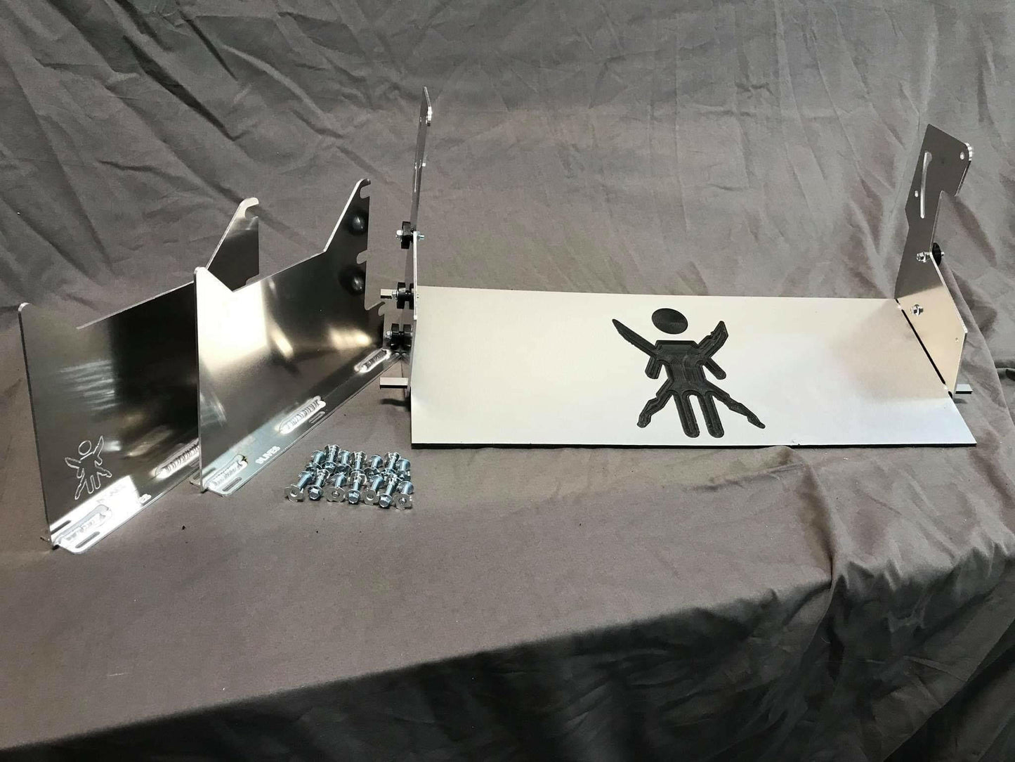 A Miata Medium Downforce Kit '90-05 NA/NB by 9livesracing on a gray surface, featuring several flat metal pieces with intricate cutouts, along with a pile of screws, nuts, and bolts, ready for assembly. The design includes a larger flat piece with a stylized human figure cutout reminiscent of an aerodynamic kit for an NB Miata.