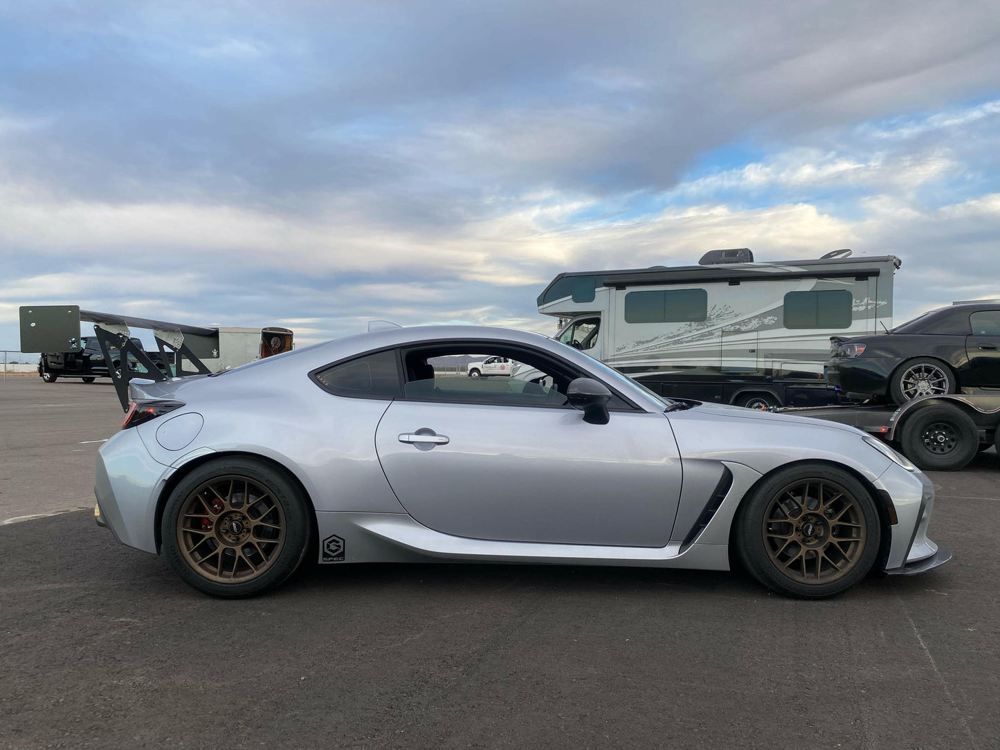 A sleek silver GR86 / BRZ fitted with bronze wheels is parked on an asphalt surface. The 9livesracing Extruded Aluminum Wings enhance its sporty appeal. In the background, there are a few other cars and a large camper under a partly cloudy sky.