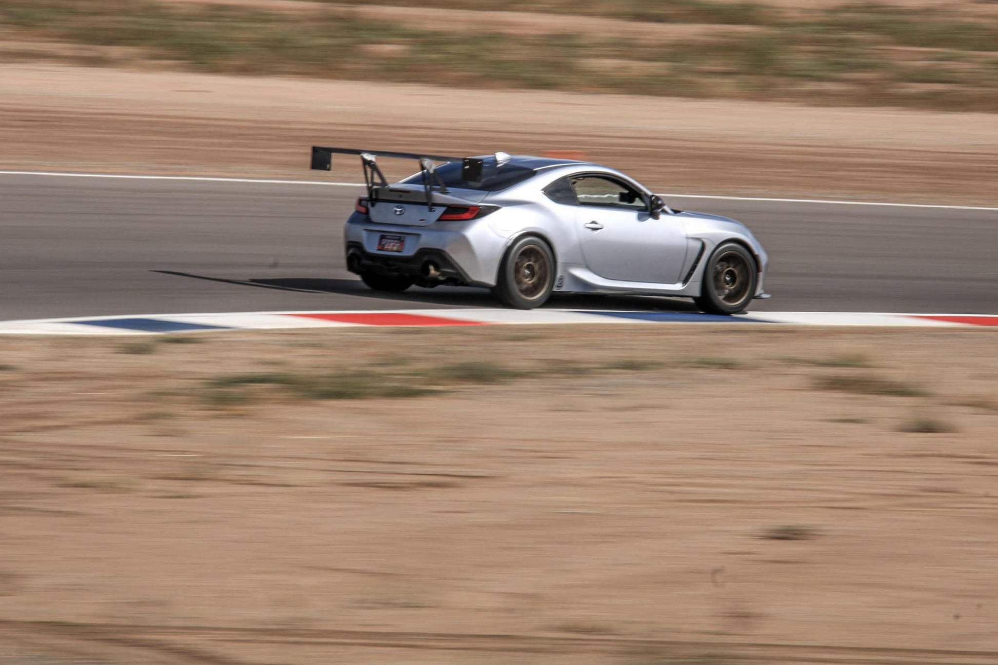 A silver sports car equipped with the 9livesracing GR86/BRZ Wang Kit '22+ and extruded aluminum wings is speeding on a racetrack. The car leans slightly as it navigates a turn, with blurred surroundings indicating high speed. The tarmac is bordered by red, white, and blue stripes, contrasting against the sandy landscape in the background.