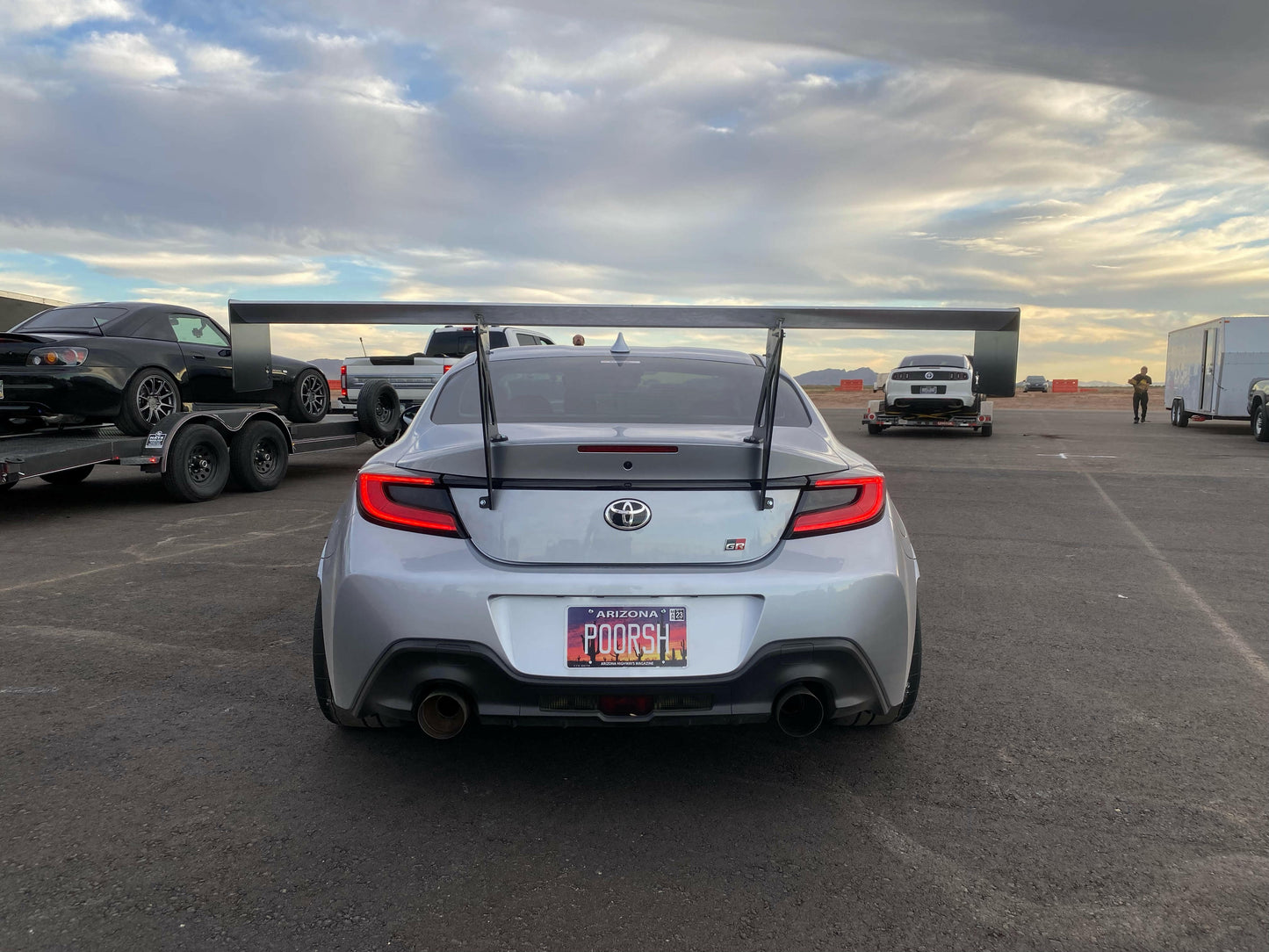 A silver Toyota sports car, equipped with the 9livesracing GR86/BRZ Wang Kit '22+ featuring large extruded aluminum wings that enhance its lift-to-drag ratio, is parked on an asphalt surface. Its license plate reads "POORSH" and displays an Arizona state emblem. In the background, another sports car is on a trailer alongside several other vehicles and a building under a cloudy sky.