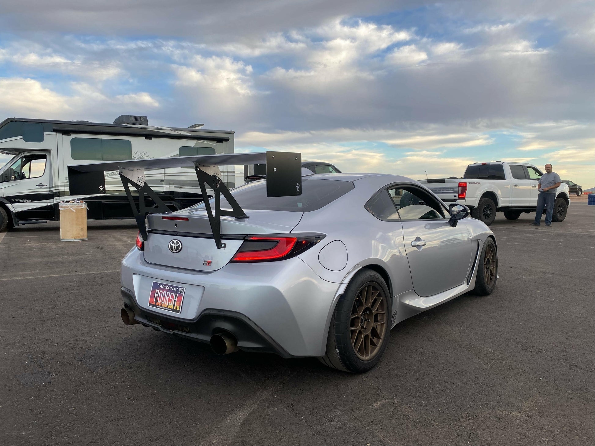 A silver Toyota sports car, equipped with a GR86 / BRZ Wang Kit '22+ from 9livesracing, featuring performance airfoils on its large rear spoiler, is parked in a lot. The car has a custom license plate reading "PROBRO." In the background under a partly cloudy sky, an RV and a white SUV are visible, along with two people standing near the SUV.