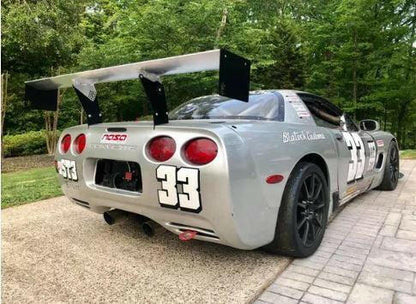 A silver Corvette race car with the number 33 displayed on its sides and back is parked on a paved driveway. The car boasts 9livesracing's Corvette C5/C6 Mounts - Pylons Only for added stability, along with a large rear spoiler, black wheels, and various decals. Trees and greenery are visible in the background.